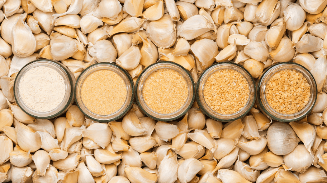 Various glass bottles filled with garlic powder behind backdrop of garlic cloves
