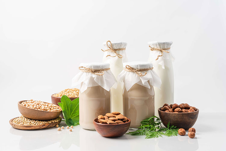 Medium shot of a four glass bottles filled with plant based milk with bowls filled with almonds, macadamia and pecan nuts.