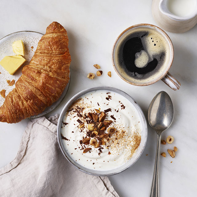 Above shot of a croissant with, yogurt sprinkled with nuts, coffee in a mug and fresh milk on the side