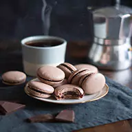 A plate filled with chocolate macarons, a cup of coffe on the side