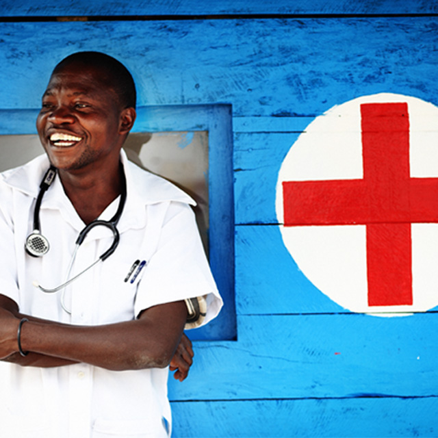 Smiling male nurse posing with his arm crossed and standing against a blue wall with a red cross on it